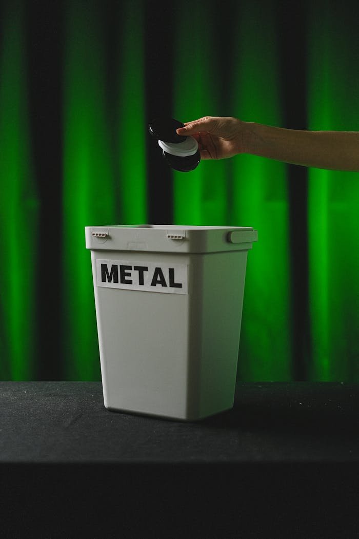 A hand placing bottle caps in a metal recycling bin against a green backdrop.